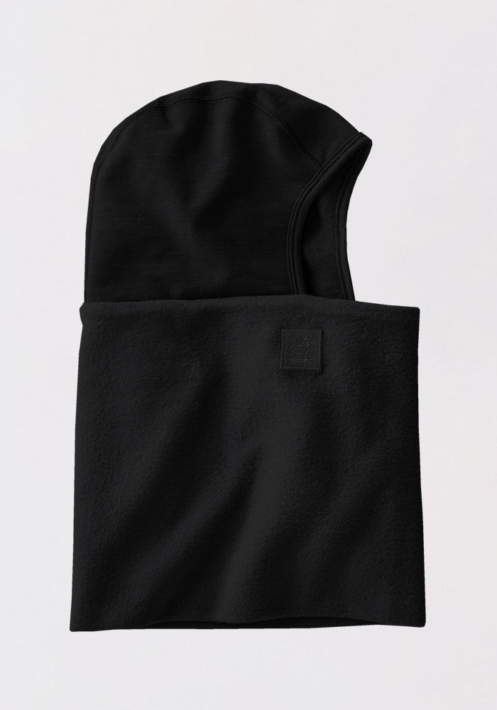 The Woolx Merinoclava in black is a soft, fleece-like balaclava with an extended neck, helmet compatibility, and a rectangular patch near the opening. Laid flat on a light background, it offers excellent cold weather protection.