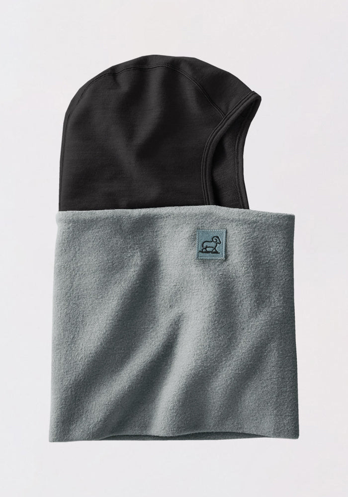 The Woolx Merinoclava in Sedona Sage features a black hood attached to a light gray fleece neck warmer with a small blue embroidered logo patch on the front. Helmet compatible for cold weather protection. Shown on a plain white background.