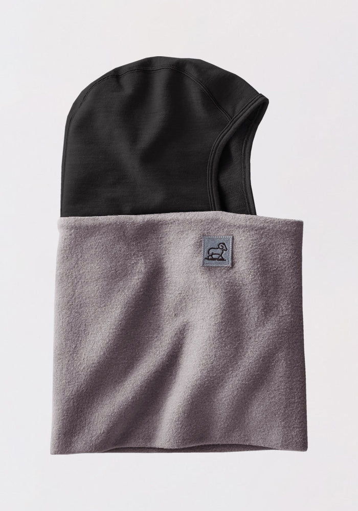 The Woolx Merinoclava in Arctic Dusk is a two-toned, helmet-compatible hooded neck warmer with a black hood, light gray fleece covering, and a small rectangular front logo patch for extra cold weather protection.