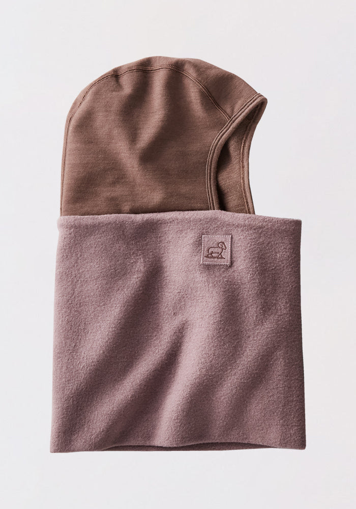 The Woolx Merinoclava in Plum Truffle is a fleece neck warmer with a built-in brown hood, neatly folded and featuring an embroidered animal patch, designed for cold weather protection.