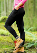 Standing outdoors on mossy ground, a person wears Woolx MerinoFlex® McKenna Pocket Leggings in black, paired with a pink long-sleeve shirt, gray socks, and tan hiking boots. A blurred forest backdrop completes the active look.