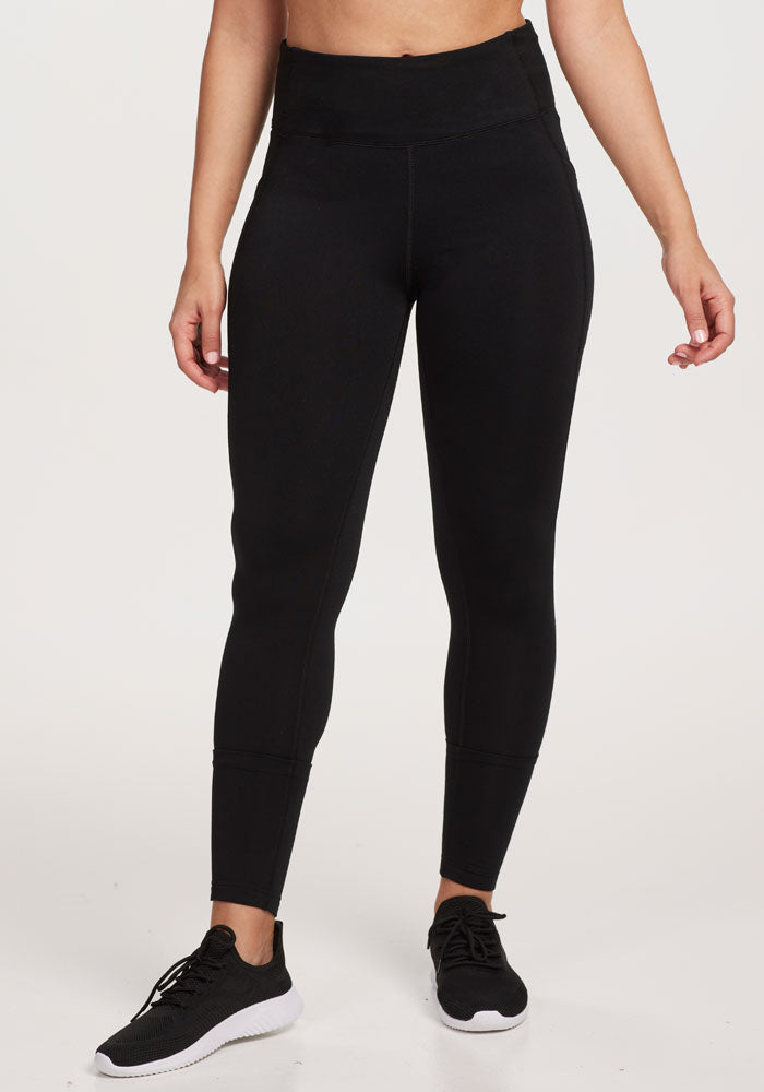 A person stands against a plain background wearing Woolx MerinoFlex® McKenna Pocket Leggings in black with side pockets and black athletic shoes; their arms are relaxed at their sides and their head is out of frame. | Tori is 5'7", wearing a size S