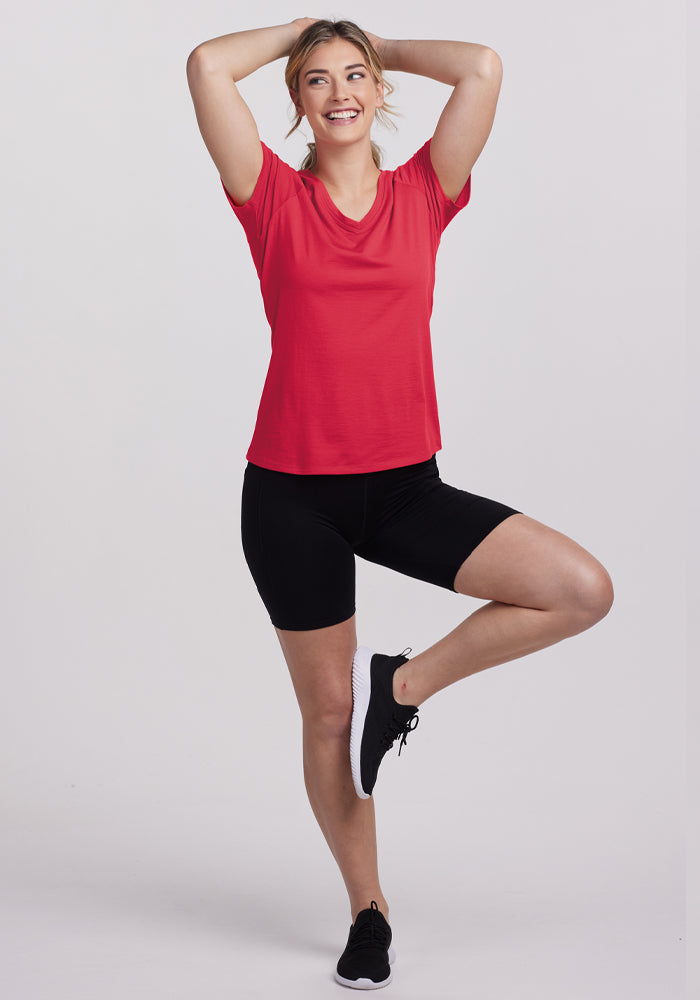 Woman wears Woolx Mia V Neck - Berry Ice Pop, smiling in a yoga pose with hands on head against a light background.