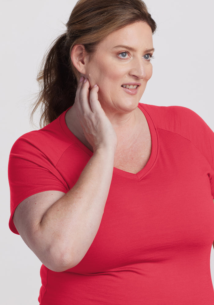 Smiling woman with brown ponytail wears Woolx Mia V Neck - Berry Ice Pop t-shirt, touching her neck, on plain background.