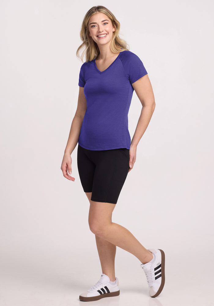 Smiling woman in a Woolx Mia V Neck - Berry Indigo tee, black shorts, and white sneakers against a light background.