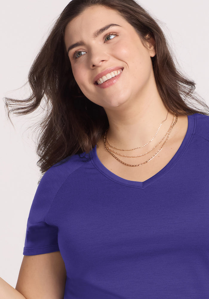 Woman with long brown hair smiles, looking up in the Woolx Mia V Neck - Berry Indigo and layered gold necklaces.