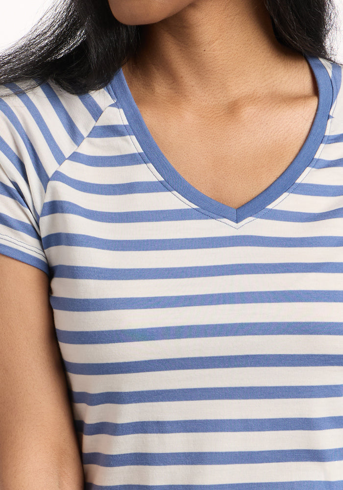 A person wears the Woolx Mia V Neck in Blue Sand Stripe—a short-sleeved, V-neck women's Merino wool tee with blue and white stripes. Only the upper body and part of the face are visible. The fabric is breathable and odor-resistant.