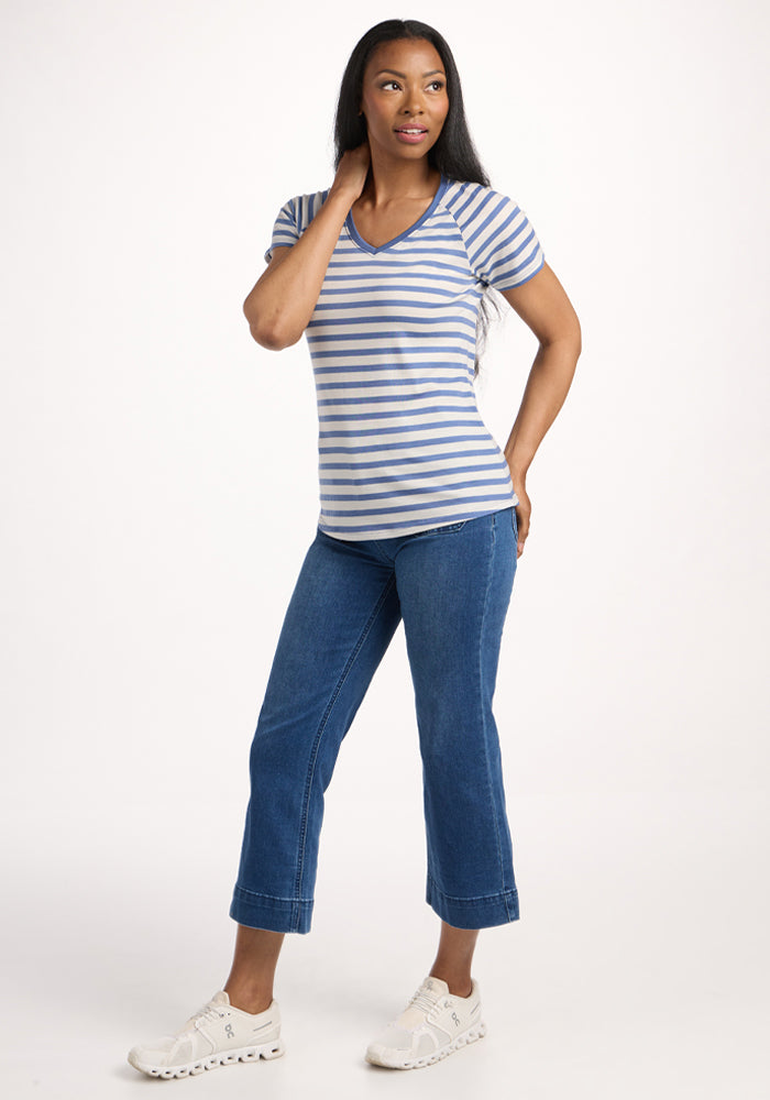 A woman with long hair poses confidently against a white background, wearing the Woolx Mia V Neck in Blue Sand Stripe, blue cropped jeans, and white sneakers.