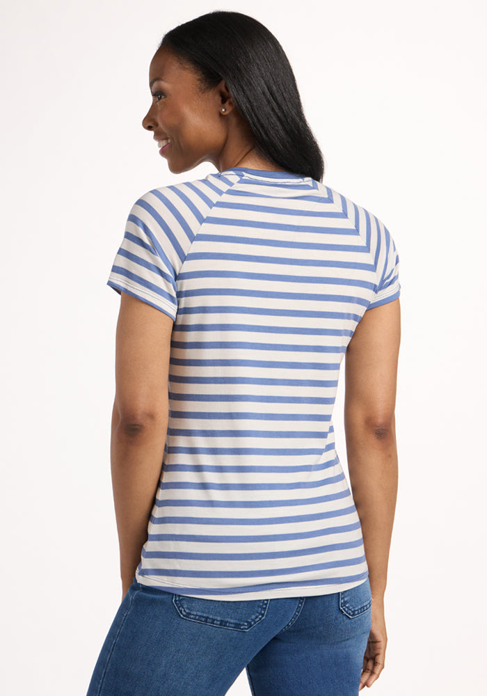 A woman with long dark hair smiles facing away, wearing the Woolx Mia V Neck in Blue Sand Stripe—an odor-resistant, moisture-wicking Merino wool tee—and blue jeans against a plain light background.