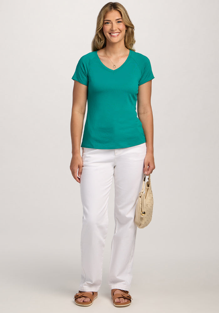 Smiling woman in a Woolx Mia V Neck - Cabana Crush tee, white pants, sandals, and beige purse stands against a plain background.