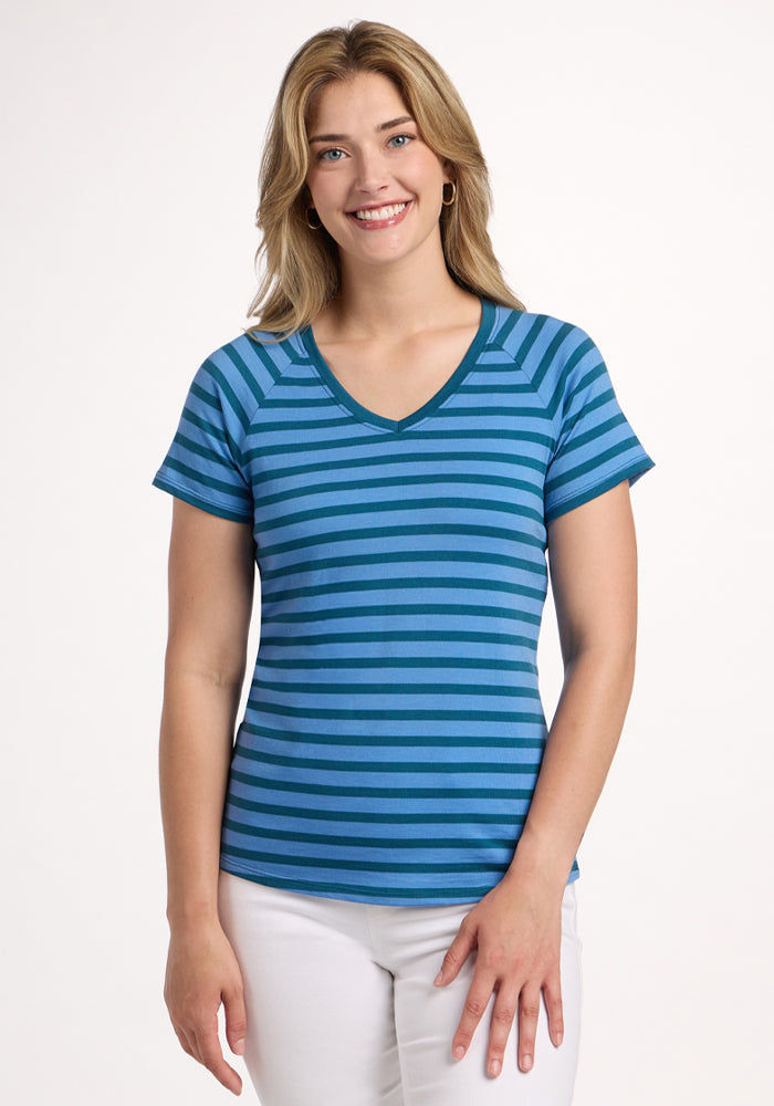 A smiling woman with shoulder-length blonde hair wears the Woolx Mia V Neck - Coastal Stripe short-sleeve shirt and white pants, standing against a plain white background. | Ariana is 5'11", wearing a size S