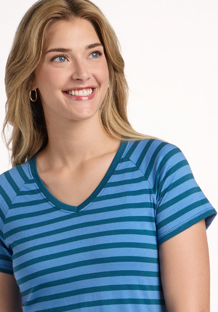 A smiling woman with long blonde hair and gold hoop earrings wears the Woolx Mia V Neck - Coastal Stripe, a women’s Merino wool tee known for odor resistance and moisture wicking, looking off to the side against a plain white background.