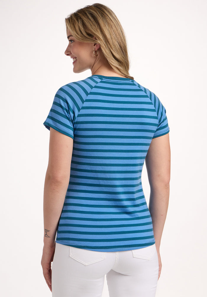 A blonde woman is shown from the back, smiling in Woolx’s Mia V Neck - Coastal Stripe tee and white pants, against a plain white background.