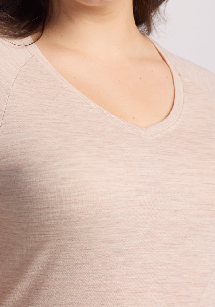 A close-up shows someone wearing the Mia V Neck in Cream Heather by Woolx. Known for its texture and fit, this Merino wool T-shirt is odor-free and offers timeless comfort. The plain, light background enhances the shirt's elegant simplicity.