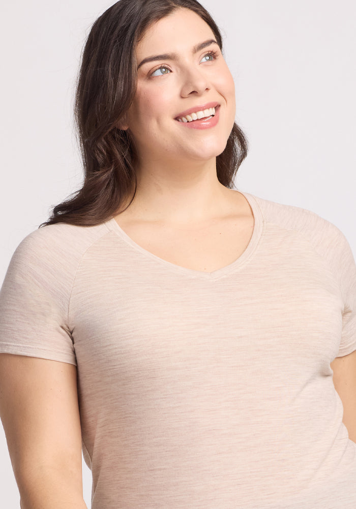 A woman with long brown hair smiles as she gazes upward and to her right. She's wearing the Woolx Mia V Neck in Cream Heather against a plain, light-colored background.