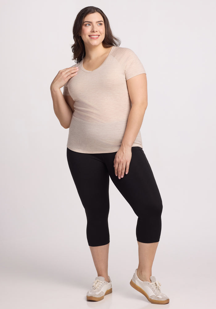 A person stands smiling against a plain background, wearing the Woolx Mia V Neck in Cream Heather, black leggings, and beige sneakers. Their shoulder-length dark hair adds charm to their pose with one hand on their hip.
