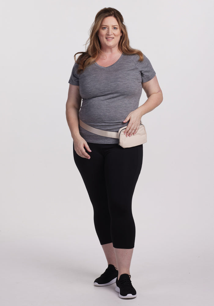 A woman poses against a white background, smiling in Woolx's Mia V Neck - Graphite Heather. She pairs it with black leggings, black sneakers, and a beige waist bag. Her long brown hair flows naturally.