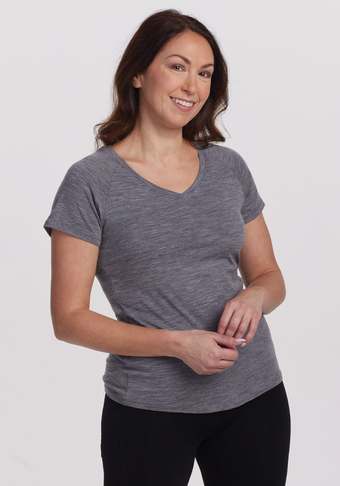 Wearing a comfortable Woolx Mia V Neck in Graphite Heather and black pants, a person smiles while standing against a plain light background, casually holding their hands together in front. | Tiffany is 5’8”, wearing a size S