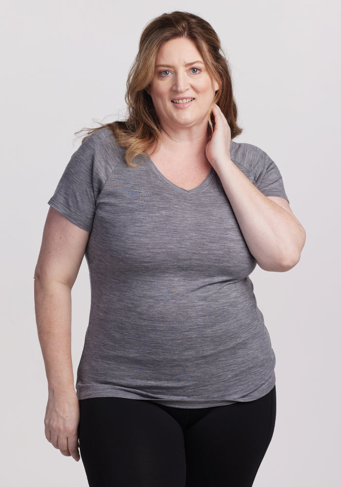 A woman with long brown hair smiles and touches her neck, wearing the comfortable Woolx Mia V Neck - Graphite Heather tee and black pants, standing against a plain white background. | Cambre is 5'11", wearing a size XL