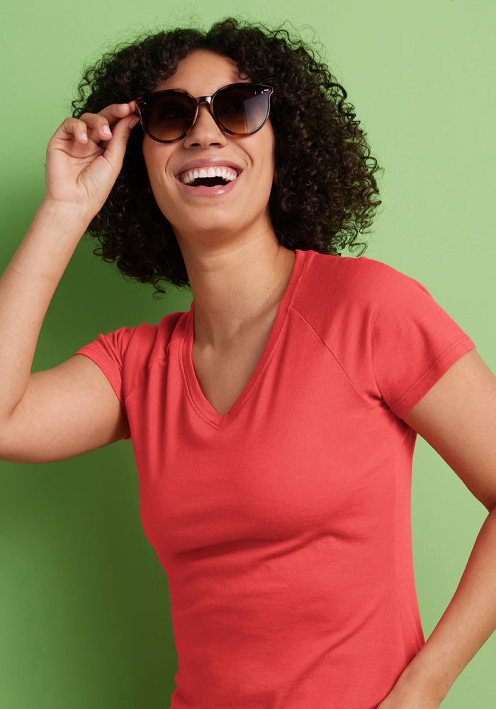 Smiling woman in a Woolx Mia V Neck - Hibiscus shirt and sunglasses stands against a green background, hand on glasses.