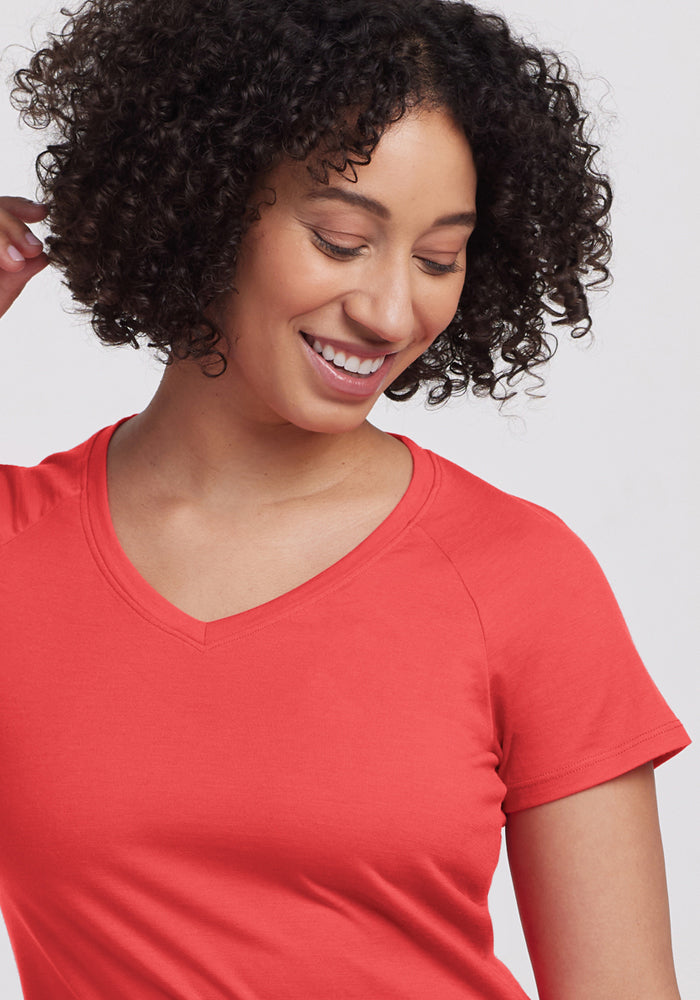 Woman with curly hair in a Woolx Mia V Neck - Hibiscus shirt smiles, looking down against a plain light background.