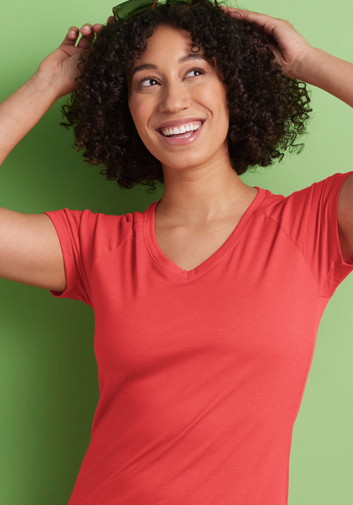 Smiling woman in Woolx's Mia V Neck - Hibiscus shirt touches her sunglasses against a green background.