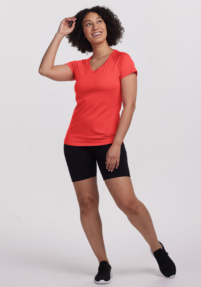 Smiling woman wears Woolx Mia V Neck - Hibiscus and black shorts, posing with one hand raised against a plain background.
