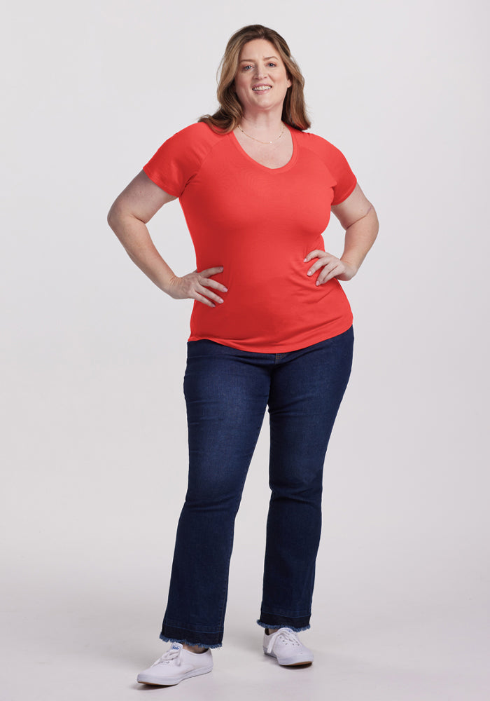 Woman smiling, wearing Woolx Mia V Neck - Hibiscus shirt, dark jeans, and white sneakers against a plain background.
