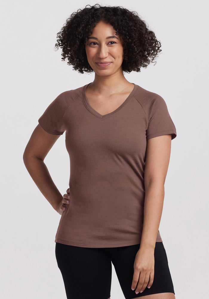 Smiling woman with curly hair wears a Woolx Mia V Neck - Peppercorn and black shorts, one hand on her hip. | Tori is 5'7", wearing a size S