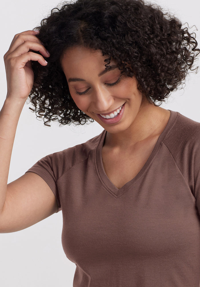 Smiling woman with curly hair touches her hair in the Woolx Mia V Neck - Peppercorn against a light background.