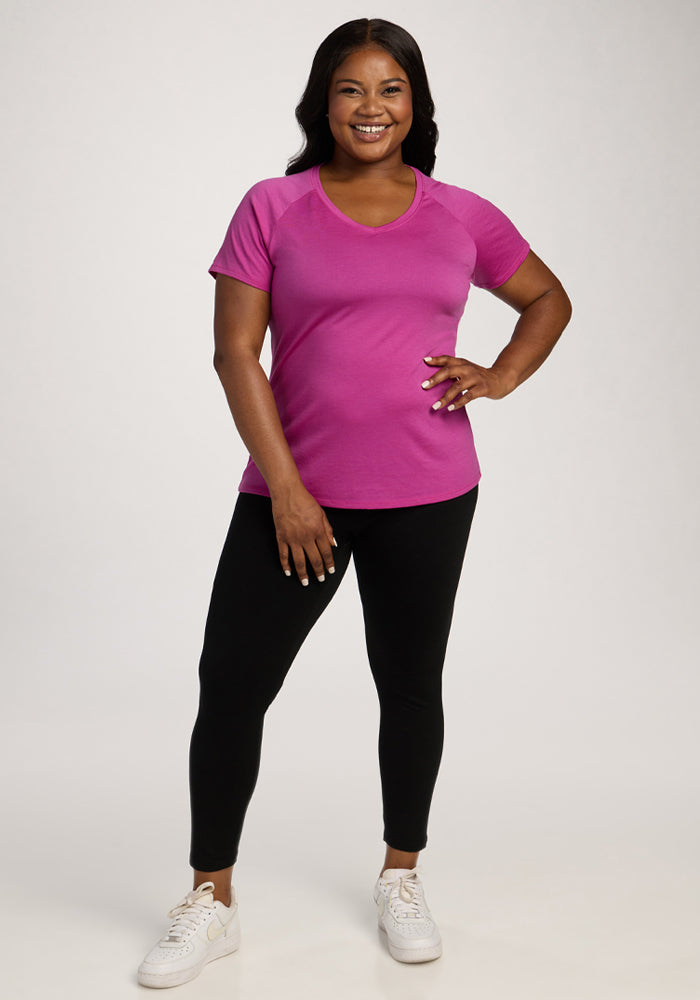Woman smiling in a Woolx Mia V Neck - Summer Sorbet shirt, black leggings, and white sneakers against a plain background.