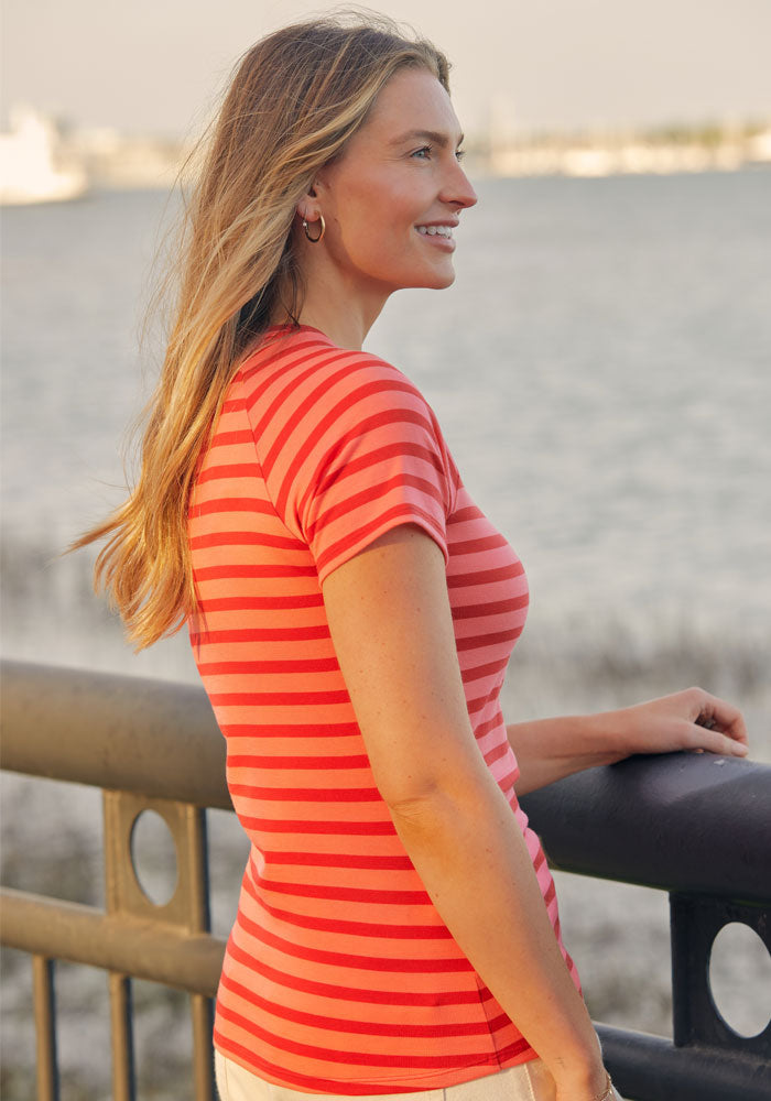 A woman with long, blonde hair smiles by a waterfront railing, wearing the Woolx Mia V Neck - Wildberry Stripe tee. The water and a blurred shoreline appear in the background.