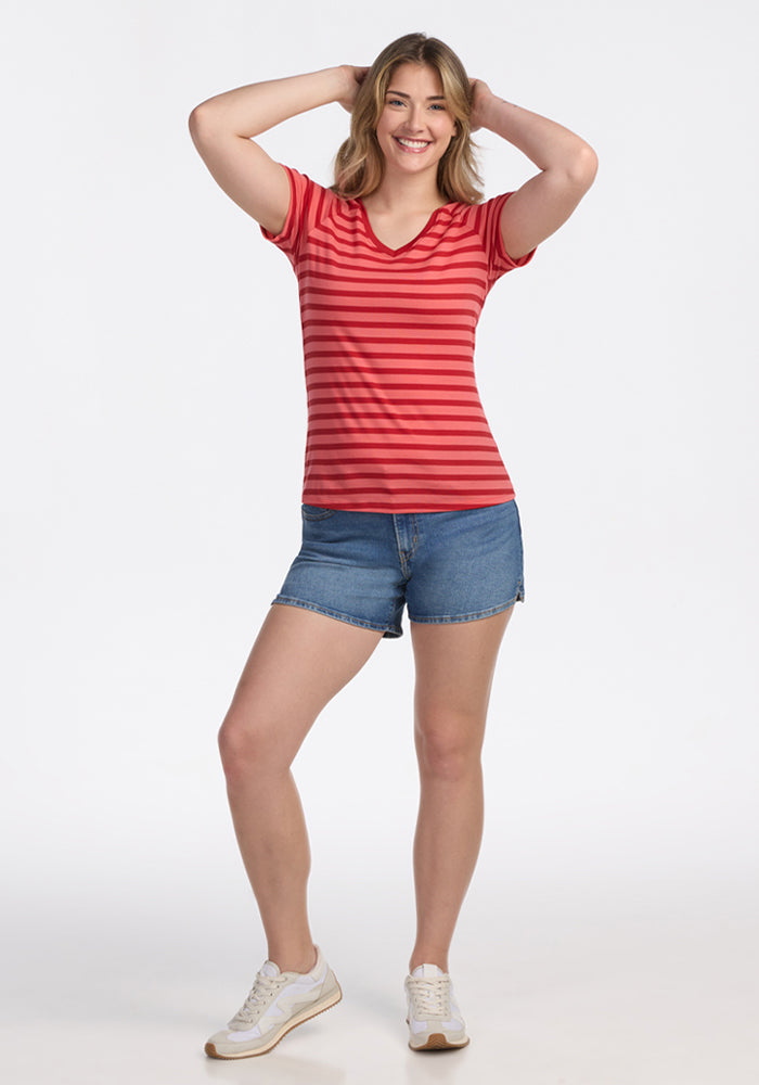 A smiling woman poses with her hands on her head, wearing the Woolx Mia V Neck in Wildberry Stripe, denim shorts, and white sneakers against a plain white background.