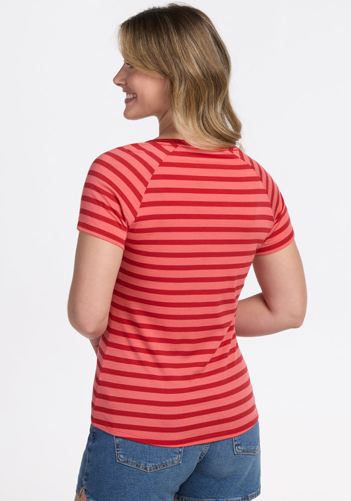A woman with shoulder-length blonde hair smiles and looks to the side, wearing the Woolx Mia V Neck in Wildberry Stripe and denim shorts, standing against a plain white background.