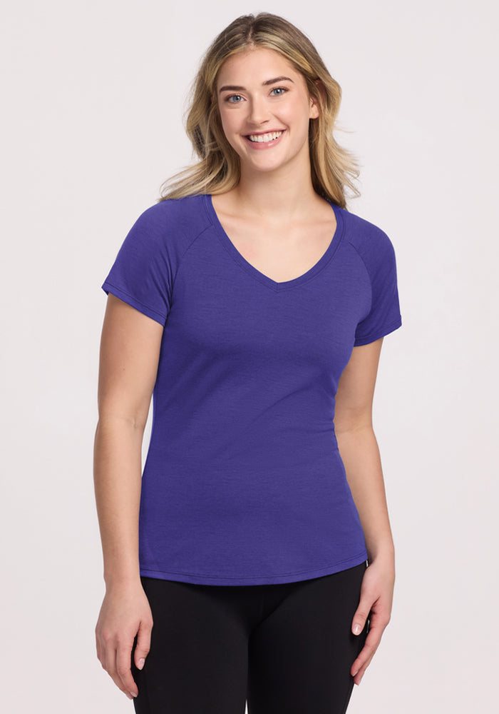 Smiling woman with long blonde hair wears the Woolx Mia V Neck - Berry Indigo tee and black pants against a light background. | Ariana is 5'11", wearing a size S