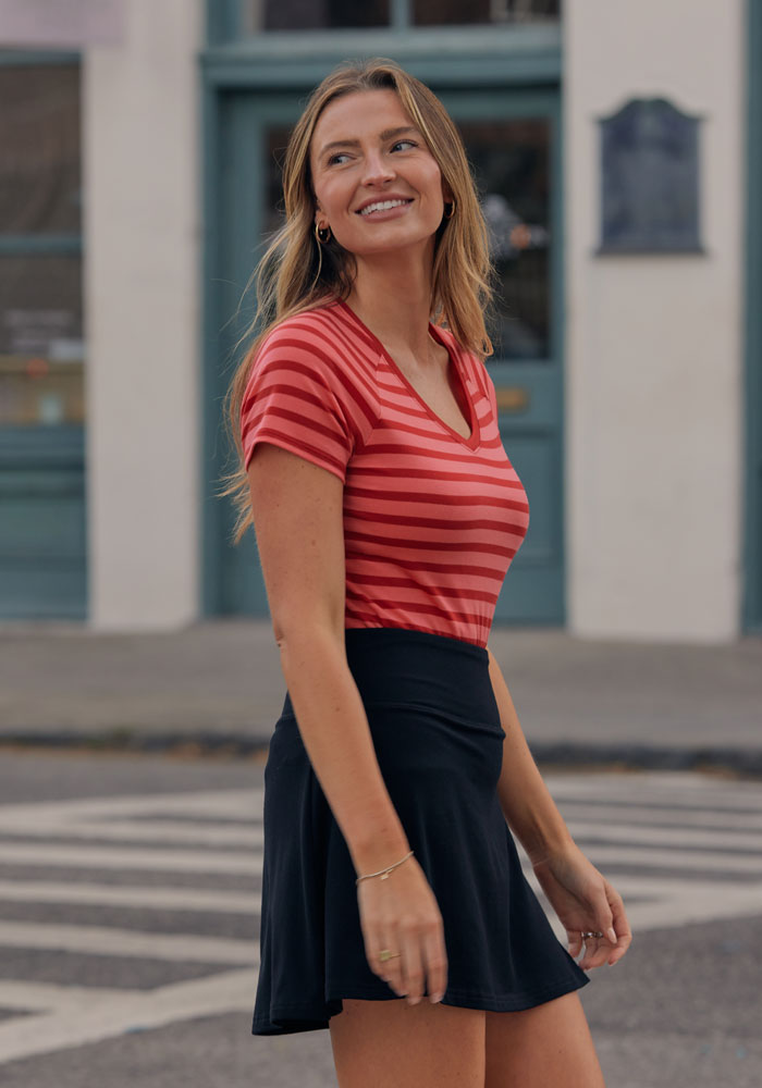 A woman with long, light brown hair smiles outdoors in the Woolx Mia V Neck - Wildberry Stripe, paired with a black skirt. Blue windows and doors in the background highlight her comfortable, stylish look.