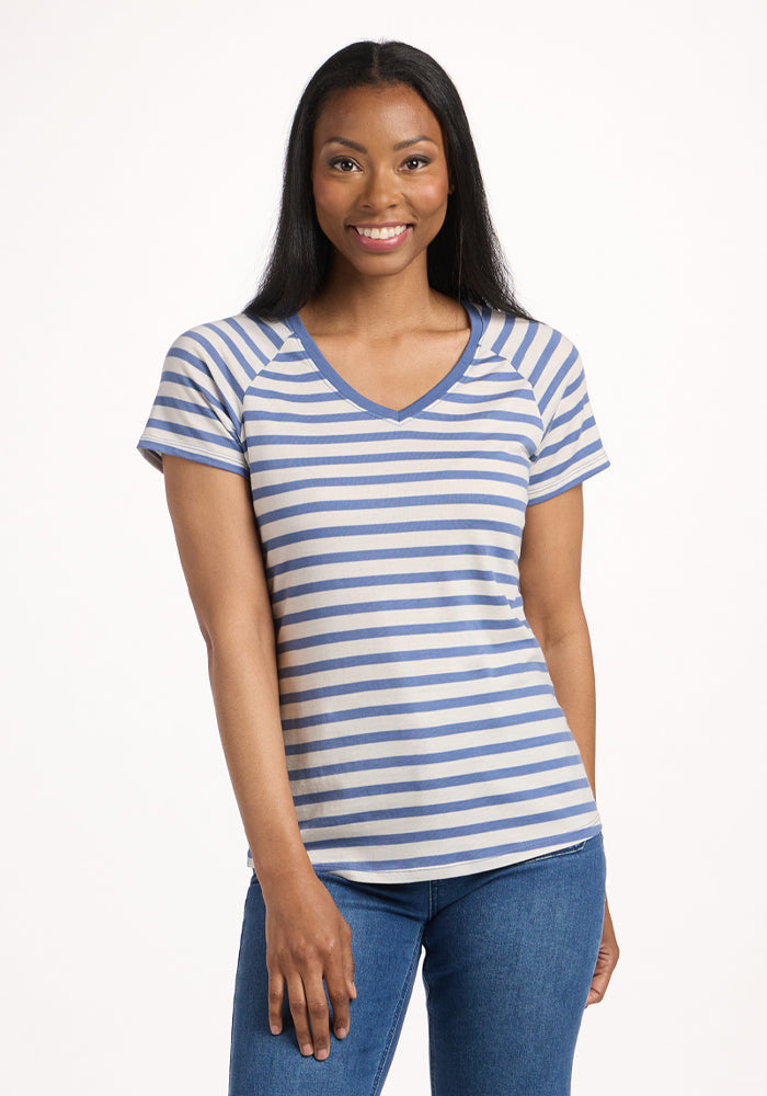 A woman with long dark hair smiles in a Woolx Mia V Neck - Blue Sand Stripe short-sleeved, moisture-wicking shirt and blue jeans, standing against a plain white background. | Jade is 5’10”, wearing a size S