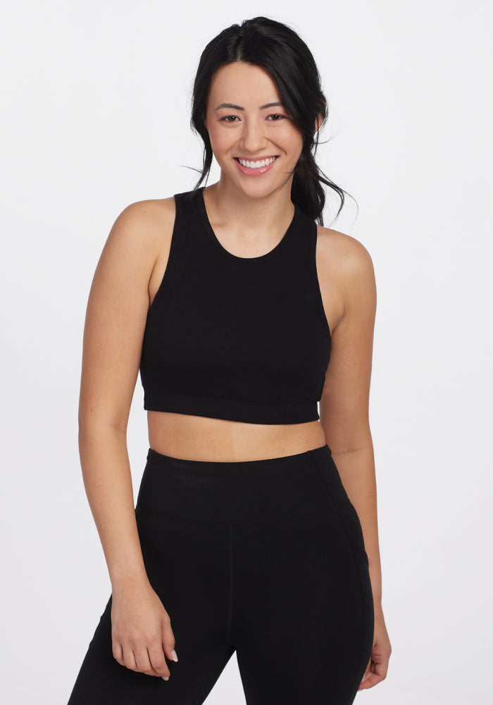 A person with long dark hair smiles, wearing Woolx's Black Mila Bra along with a black sleeveless crop top and matching high-waisted athletic pants, standing against a plain white background. | Sarah is 5'8", wearing a size S