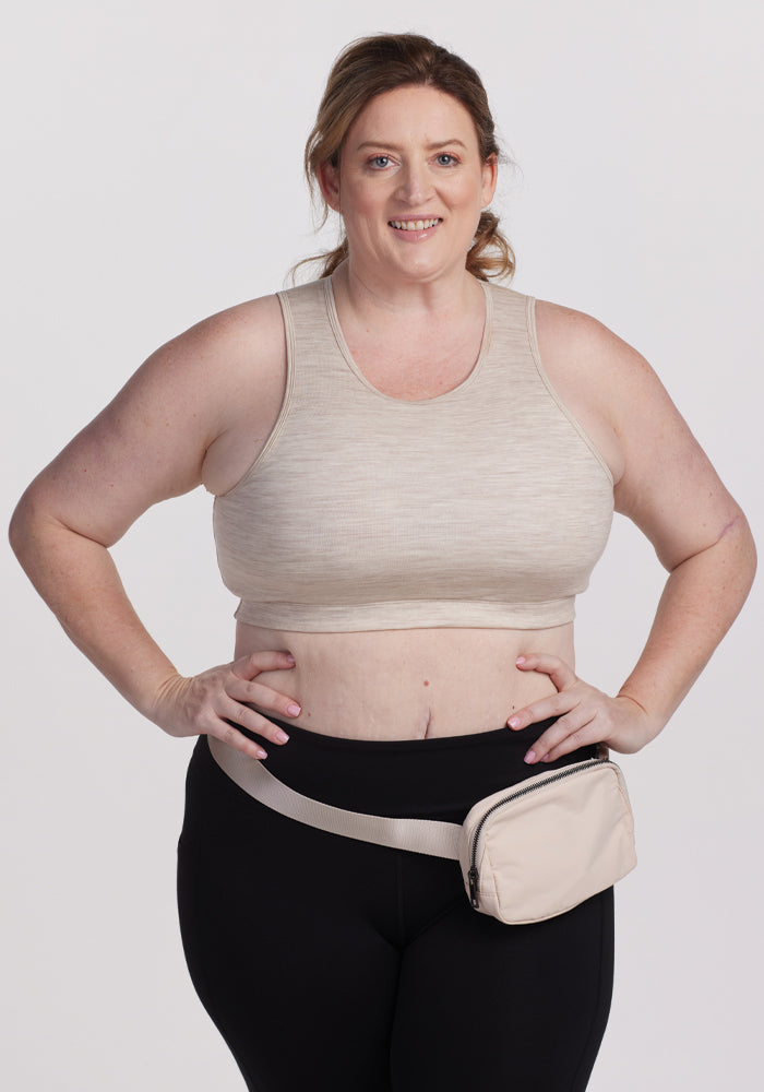 A person wearing a Woolx Mila Bra in Cream Heather smiles at the camera, paired with black leggings and holding a matching beige waist pack, set against a plain, light-colored backdrop. | Cambre is 5'11", wearing a size XL