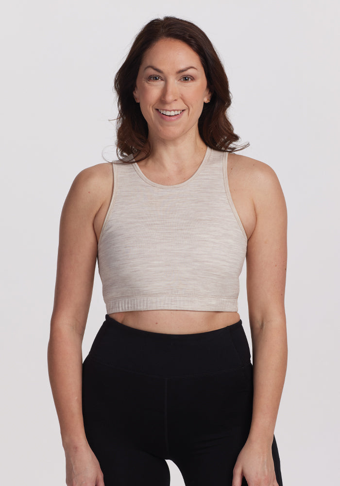 A person with long brown hair, wearing a beige sleeveless top and black leggings, smiles confidently against a white background in a comfortable Woolx Mila Bra - Cream Heather. | Tiffany is 5’8”, wearing a size S