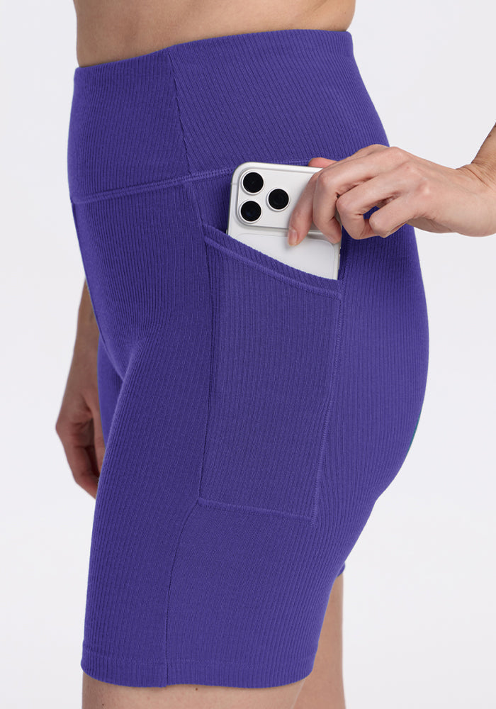 Person sliding a smartphone into the side pocket of Woolx Miley Bike Shorts - Berry Indigo—workout shorts with pockets.