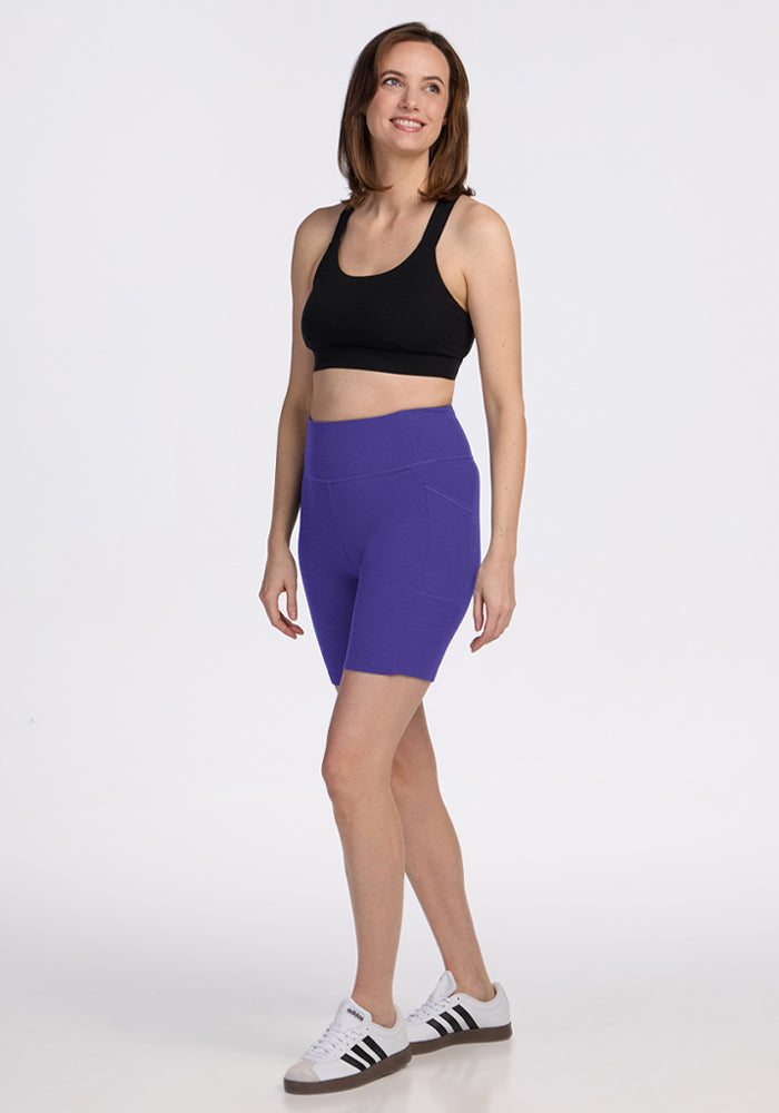 Woman wears Woolx Miley Bike Shorts in Berry Indigo, a black sports bra, and white sneakers, smiling against a plain background.