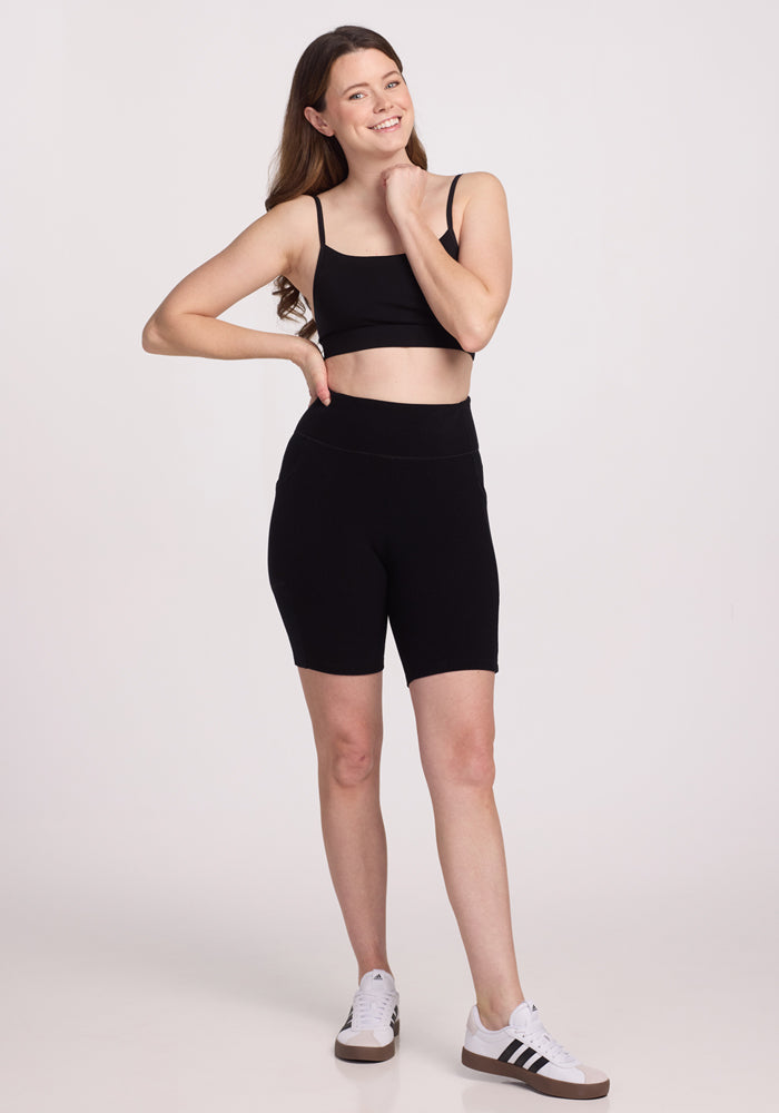 A woman in a black sports bra and Miley Bike Shorts - Black by Woolx stands smiling with one hand on her hip, wearing white sneakers with black stripes against a plain white background.