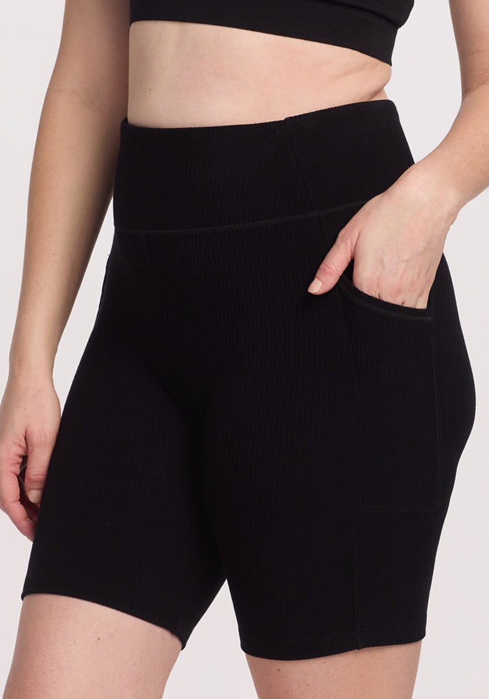 A person stands against a plain background wearing Woolx's Miley Bike Shorts - Black, high-waisted with a side pocket, paired with a black crop top.