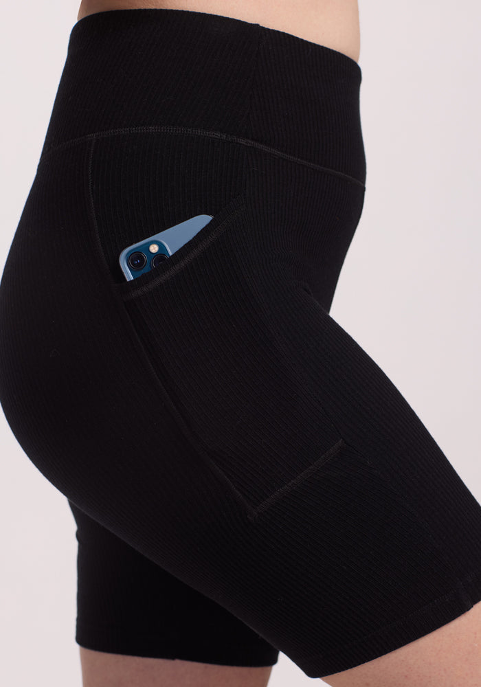 A close-up shows a person wearing Woolx Miley Bike Shorts in black, with a smartphone peeking from the side pocket against a neutral background, highlighting the shorts' ribbed texture and design.