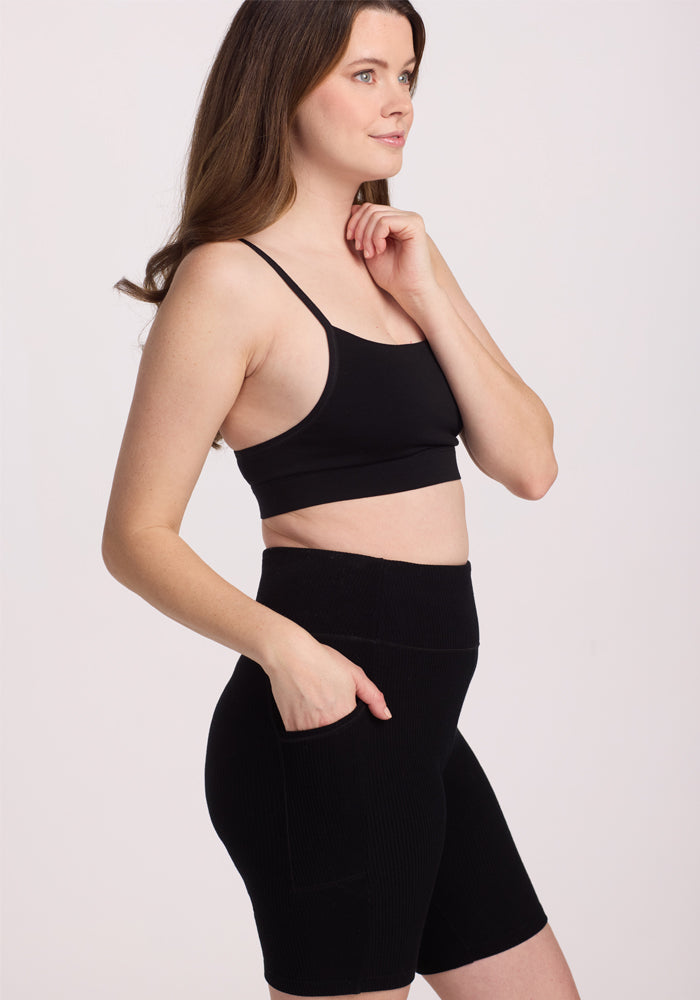 A woman with long hair stands in profile, wearing a black sports bra and Woolx Miley Bike Shorts. One hand rests on her chin, the other by her side against a plain white backdrop.