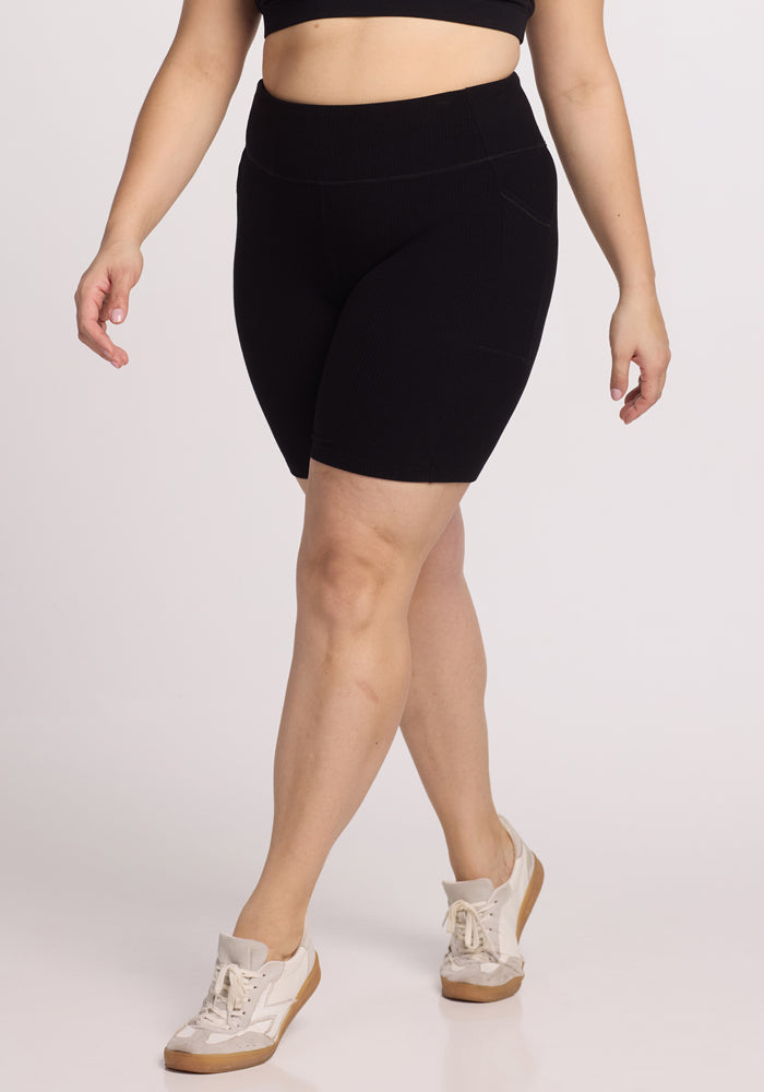 A person in Woolx's Miley Bike Shorts - Black and a matching sports bra stands against a plain background, wearing white sneakers with light tan soles, casually stepping forward. | Juliana is 5’10”, wearing a size XL
