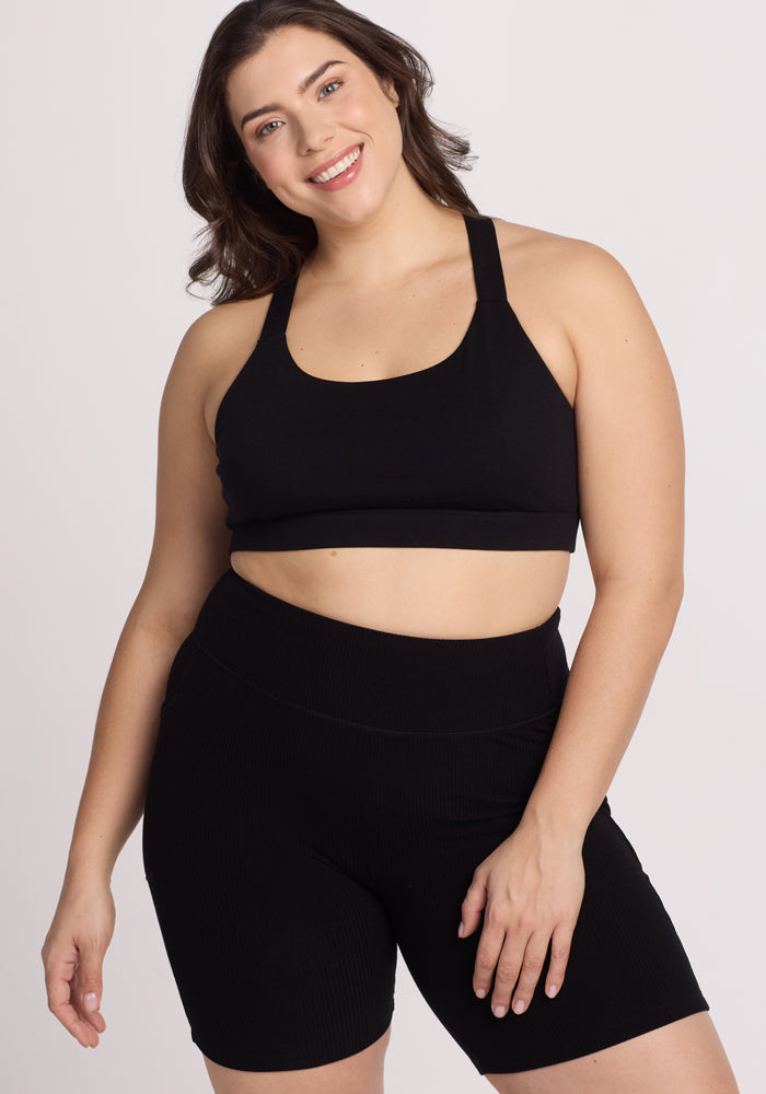 A woman smiling and posing confidently in a Woolx black sports bra and Miley Bike Shorts against a plain white background.