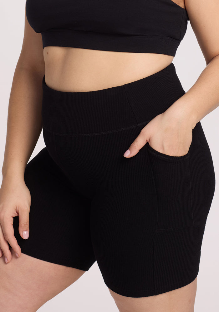 A person wearing a black sports bra and Woolx's Miley Bike Shorts - Black. The image highlights the midsection and legs, showcasing the texture and fit of the ribbed shorts with pockets against a plain white background.