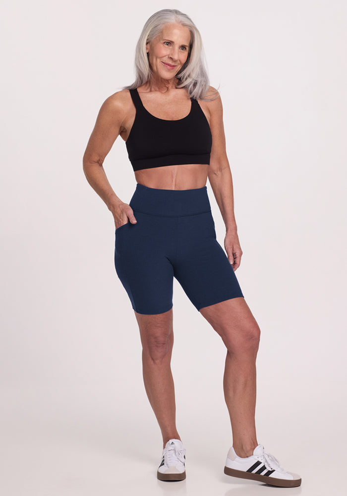 Smiling older woman wears Woolx Miley Bike Shorts in Deep Navy, black sports bra, and white sneakers, hand in pocket.